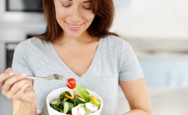 Mujer sonriente disfrutando de una ensalada fresca y saludable, con ingredientes como tomate, lechuga y champiñones, representando la conexión entre alimentación saludable y bienestar.