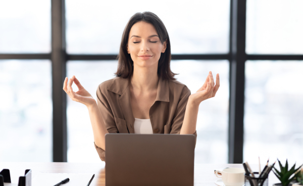 Mujer joven practicando meditación frente a su computadora en una oficina, con expresión relajada y gesto de yoga.